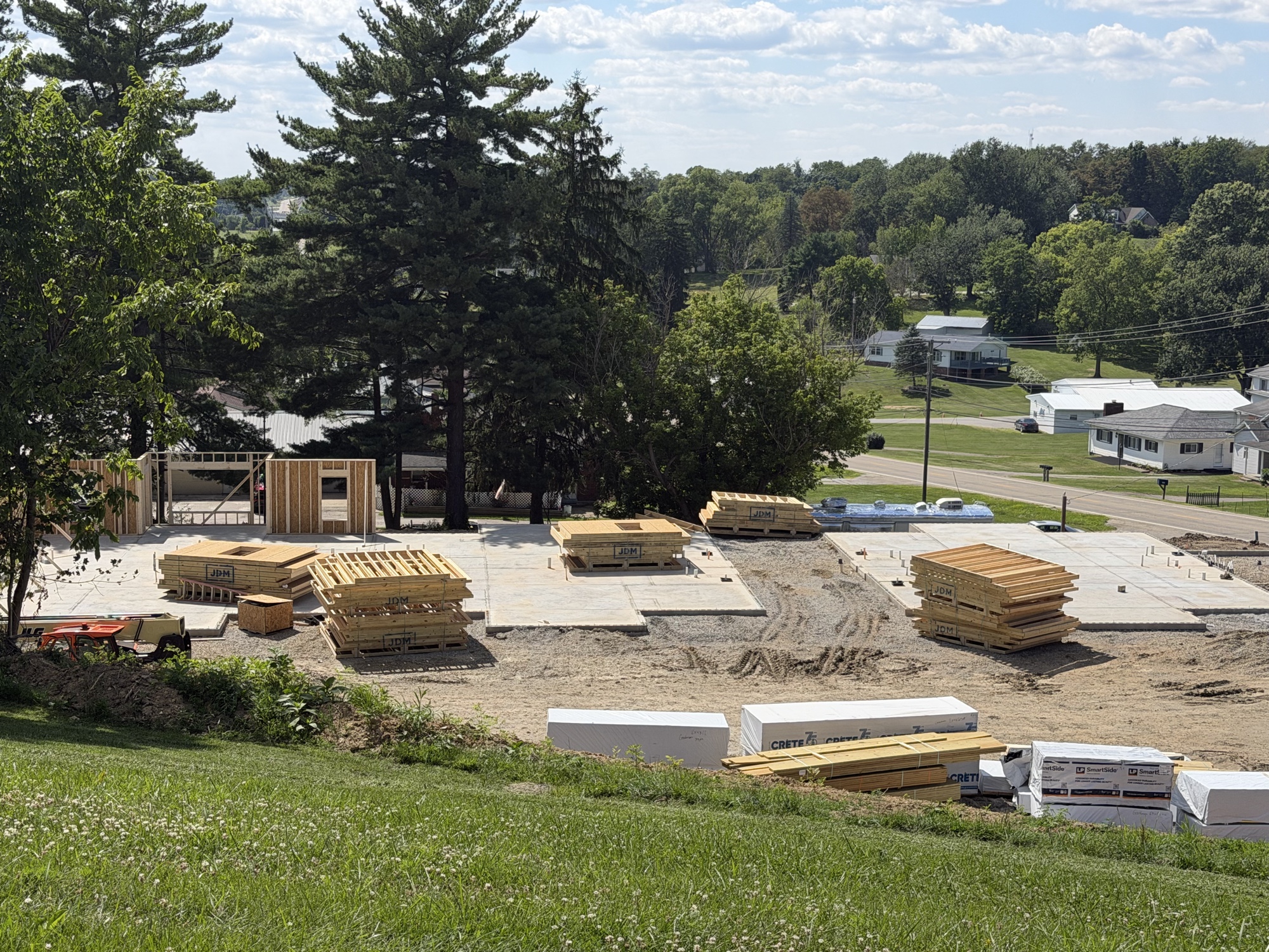 Real construction photo: first exterior wall section standing on a completed foundation slab at The Legacy at Richland Commons, with prefabricated floor truss bundles staged nearby, July 30, 2026